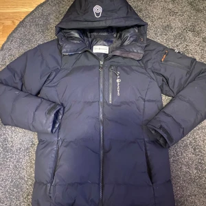 Svart dunjacka från Sail Racing - Säljer en svart dunjacka från Sail Racing med huva och dragkedja. Jackan har en cool expedition-patch på ärmen och är perfekt för kalla dagar. Tillverkad i nylon med dunfyllning för extra värme. Passar bra för vinteräventyr och håller dig varm och bekväm.