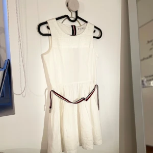 vit klänning-Tommy Hilfiger - Snygg vit ärmlös klänning från Tommy Hilfiger med ett stilrent bälte i rött, vitt och blått. Klänningen har en dragkedja baktill och är perfekt för en somrig look. Den är tillverkad i ett lätt och bekvämt material. Helt oanvänd💞