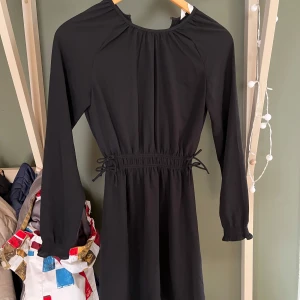 Svart klänning från H&M - Elegant svart klänning från H&M med långa ärmar. Klänningen har en elastisk midja med knytband för en justerbar passform. Perfekt för en stilren look.