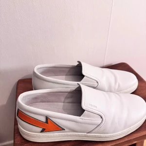 Vita slip-ons från Prada med orange detalj - Snygga vita loafers från Prada i skinn med en cool orange pil-detalj på sidan. Perfekta för en stilren look. Skorna har en diskret Prada-logga och är lätta att ta på.
