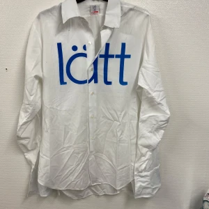 Vit skjorta med blå text - Säljer en vit skjorta med blå text 'lätt' tryckt på framsidan. Skjortan har långa ärmar och knappar framtill. Perfekt för en avslappnad stil.