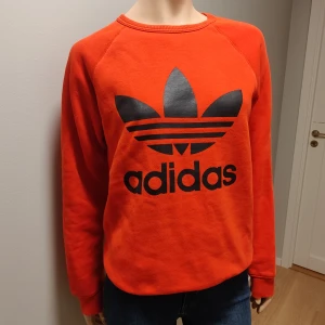 Röd  sweatshirt från Adidas - Snygg röd (ser mer orange ut på bilderna) sweatshirt från Adidas med det klassiska logotyptrycket i svart på framsidan. Tröjan har långa ärmar och en rund halsringning. Perfekt för en sportig och avslappnad stil.