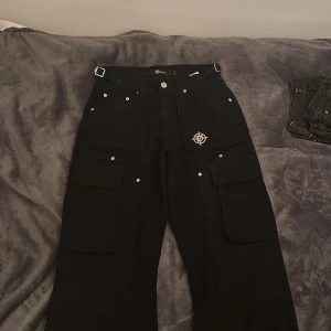 Svarta cargobyxor från Vicinity - Snygga svarta cargobyxor från Vicinity med flera fickor och en cool logga på benet. Byxorna har en loose passform och är perfekta för en avslappnad stil. De har knappar och en dragkedja framtill.