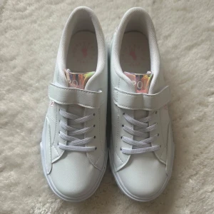 Vita sneakers från Ralph Lauren, Polo - Vita sneakers från Polo,  Ralph Lauren. Snörning och kardborreband för extra komfort. Skorna har en glansig detalj på hälen och rosa logotype på sidan och på sulan i skon. Helt nya och oanvända. 