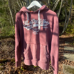 Vinröd hoodie från Superdry - Säljer en extremt snygg vinröd hoodie från Superdry med vit text och japanska tecken på framsidan. Den har en klassisk känguruficka och justerbar huva med vita snören. Perfekt för en avslappnad stil. Extremt bra skick och sticker ut från majoriteten.