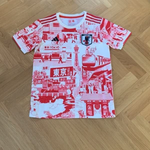 Japan kit special edition  - Säljer en unik vit och röd fotbollströja från Adidas med ett detaljerat Tokyo-motiv från Japans landslag. Tröjan har korta ärmar och tre ränder på axlarna. Perfekt för fotbollsfans som vill ha något speciellt! 🗼⚽