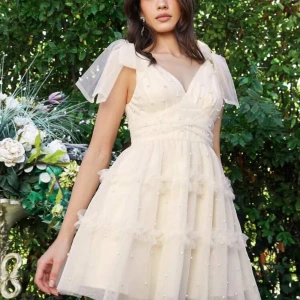 Champagnefärgad klänning med pärlor - Charmig champagnefärgad klänning med pärlor och volanger. Den har en v-ringad design och är ärmlös med tunna axelband. Perfekt för en somrig look.