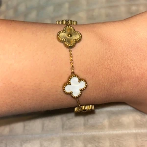 Guldarmband med fyrklöverdesign - Elegant guldarmband med fyrklöverformade länkar. Varje länk har en vit inläggning som ger en fin kontrast mot guldfärgen. Perfekt för att lägga till en touch av lyx till din stil. Guldpläterad!