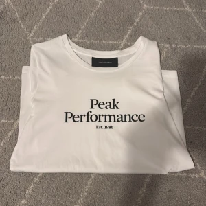 Vit t-shirt från Peak Performance - Säljer en stilren vit t-shirt från Peak Performance med tryck 'Peak Performance Est. 1986' på framsidan. Perfekt för en casual look. T-shirten är kortärmad och gjord i mjukt bomullsmaterial. Den är aldrig använd och är model dam.