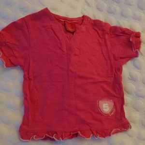 Rosa t-shirt från Esprit  storlek 74 , 6-9 månader - Söt rosa t-shirt från Esprit med texten 'Soccer Queen' och siffran 5 på baksidan. Den har korta ärmar med volangdetaljer och en liten v-ringning. Perfekt för små fotbollsfans!