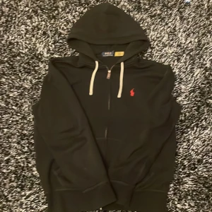 Svart hoodie från Polo Ralph Lauren - Snygg svart hoodie från Polo Ralph Lauren med dragkedja och huva. Den har en röd broderad logga på bröstet och vita snören vid huvan. Perfekt för en avslappnad stil. Köpt från Zalando för 2200 säljer för 1200 