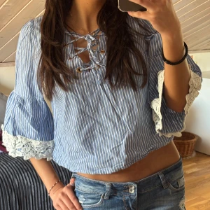 Blå- och vitrandig blus med spetsdetaljer - Säljer en söt blus i blått och vitt med randigt mönster. Blusen har trekvartsärmar med vackra spetsdetaljer och en knytning framtill. Perfekt för en avslappnad stil.