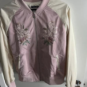 Rosa bomberjacka från Crocker - Säljer en snygg rosa bomberjacka från Crocker med vackra blombroderier på fram- och baksidan. Jackan har långa ärmar i en ljusare nyans och dragkedja framtill. Perfekt för att ge din outfit en unik touch! 🌸