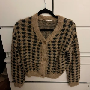 Beige och svart kofta med knappar - Mysig och fluffig kofta i beige och svart med ett rutigt mönster. Koftan har långa ärmar och knäpps med stora knappar framtill. Perfekt för kyliga dagar!