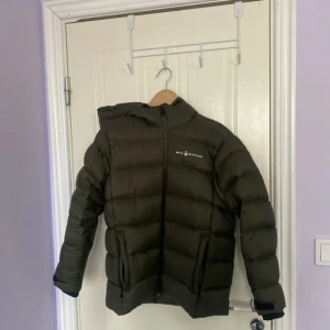 Olivgrön dunjacka från Sail Racing - Säljer en olivgrön dunjacka från Sail Racing. Jackan har en dragkedja framtill och en huva med en vit logotyp på baksidan. Perfekt för kyliga dagar med sin vadderade design.