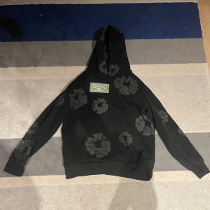 Svart hoodie från Denim Tears - Snygg svart hoodie från Denim Tears med unikt blommönster. Hoodien har en klassisk passform med långa ärmar och en praktisk magficka. Tillverkad i USA, perfekt för en stilren look.