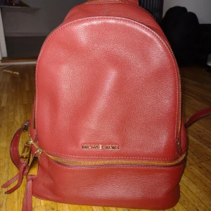 Röd ryggsäck från Michael Kors - Snygg röd ryggsäck i skinn från Michael Kors med guldfärgade dragkedjor och logotyp. Perfekt för att bära dina dagliga nödvändigheter med stil. Insidan har ett ljust foder med logotypmönster och flera fack för organisering.