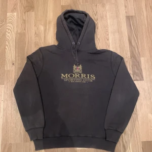 Grön hoodie från Morris - Säljer en stilren olivgrön hoodie från Morris med tryck på framsidan. Hoodien har en klassisk passform med långa ärmar och en huva.