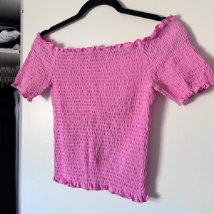 Rosa offshoulder topp - Säljer en söt rosa offshoulder topp med smockdetaljer och volangkant. Perfekt för sommarens varma dagar. Toppen har korta ärmar och en elastisk passform som ger en bekväm känsla. Passar perfekt till både jeans och kjol. 🌸 Önskar du veta exakta mått? Tveka inte att höra av dig!