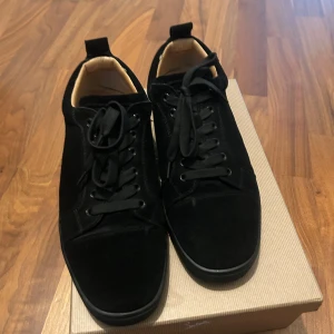 Svarta mockasneakers från Louboutin - Snygga svarta sneakers i mocka från Christian Louboutin med klassisk röd sula och svarta snören. Skorna har rund tå och platt sula, samt beige innersida för extra kontrast. Perfekta för dig som vill ha en stilren men ändå lyxig look.