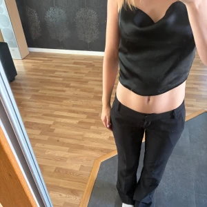 Svart satintopp med snörning - Säljer en svart croppad satintopp med tunna axelband och snygg snörning i ryggen. Toppen har en draperad urringning och glansig finish som ger en lyxig känsla. Använd en gång