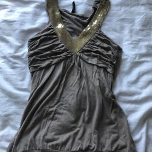 Beige linne med guldpaljetter - Snyggt beige linne med djup v-ringning och bred kant med glittriga guldpaljetter. Toppen är ärmlös och har rynkade detaljer framtill som ger en cool och trendig look. Perfekt för dig som vill sticka ut lite extra.