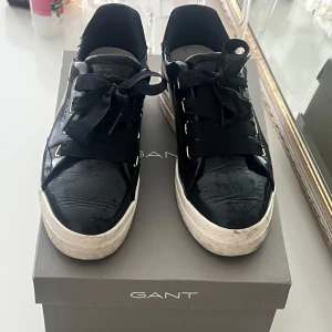 Svarta sneakers från GANT med bred snörning - Stilrena svarta sneakers från GANT med breda svarta snören och guldfärgade metalldetaljer. Skorna har en vit, chunky platt sula och är tillverkade i glansigt skinn. Perfekta för dig som gillar en clean och modern look.