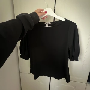 Svart blus med puffärm från H&M - En svart blus från H&M i storlek S. Toppen har rund halsringning och puffiga, korta ärmar som ger en trendig siluett. Perfekt att styla med både jeans och kjol för en clean look.