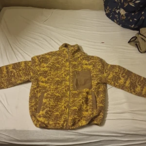 Gul och brun teddyjacka med dragkedja - Säljer en riktigt mysig teddyjacka i gul och brun mix. Jackan har hel dragkedja, hög krage och två fickor framtill med snygga detaljer. På bröstet finns en extra ficka med dragkedja. Perfekt för dig som vill ha en varm och cool look. 