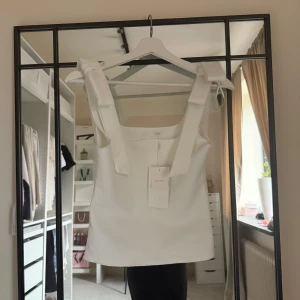 Vit linnetopp med rosettband Zara - Superfin vit linnetopp från Zara med breda axelband som knyts till rosetter på axlarna. Toppen har rak passform och ärmlös design, perfekt för en fräsch och stilren look. Materialet är mjukt och lätt, vilket gör den skön att bära under varma dagar.