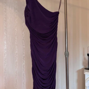 Lila one shoulder (fest)klänning H&M - Säljer en snygg lila glansig klänning från H&M med draperad design och one shoulder-modell. Klänningen har smal axelrem och är figurnära med snygga rynkade detaljer längs hela plagget. Perfekt för dig som vill sticka ut med en cool och elegant look.
