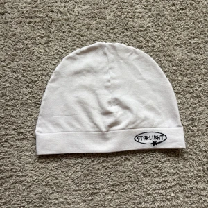 Vit mössa med Starlight-tryck - Vit, mjuk och stretchig mössa från Starlight med svart broderad logga och stjärna framtill. Enkel och clean design med uppvikt kant, perfekt för dig som gillar minimalistisk stil. Tillverkad i bomull för en skön känsla på huvudet.