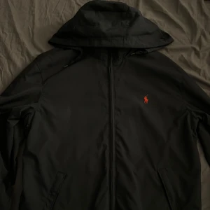 Svart vindjacka från Polo Ralph Lauren - Svart stilren och unik vindjacka från Polo Ralph Lauren med huva och dragkedja. Man kan stoppa ner huvan med en dragkedja bakomtill nacken. Insidan av kragen är fodrad för extra komfort. Perfekt för dig som vill ha en clean och stilren look. Storlek M men funkar för S som mig. 