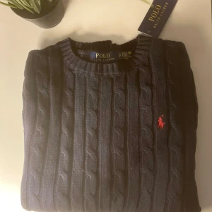 Mörkblå kabelstickad tröja Polo Ralph Lauren - Kabelstickad mörkblå tröja från Polo Ralph Lauren med klassisk rund hals och röd broderad logga på bröstet. Långärmad modell i mjuk bomull, perfekt för lager-på-lager och snygg till jeans eller kjol. Nytt skick🔥