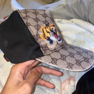 Gucci keps med tiger och GG-mönster - Snygg keps från Gucci med klassiskt GG-monogram i beige och brunt, broderad tiger framtill och svart mesh på sidorna och bak. Justerbar rem med gröna och röda ränder. Perfekt statement-piece för dig som vill sticka ut.