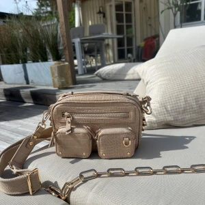 Beige axelväska med krokoprägling - Snygg beige axelväska med krokopräglat mönster och guldfärgade detaljer. Väskan har flera fack med dragkedja och två små avtagbara fickor framtill. Bred justerbar axelrem i textil och kedjedetalj. Perfekt för dig som vill ha både stil och funktion.