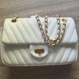 Vit quiltad axelväska från Gina Tricot - Priset kan diskuteras!    Supersnygg vit axelväska från Gina Tricot med quiltad design och guldfärgad kedja. Väskan har ett klassiskt lock med vridspänne och kedjan är flätad med vitt material. Perfekt storlek för det viktigaste och ger en clean look till din outfit.