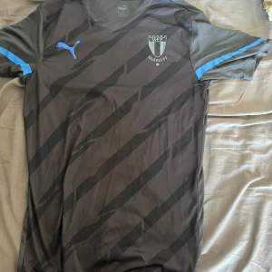 Malmö FF Tröja  - Snygg svart fotbollströja från Puma med Malmö FF-logga på bröstet och blå Puma-logga. Tröjan har svarta diagonala ränder och blå detalj på ärmarna. Perfekt för dig som vill visa ditt stöd för MFF på match eller träning. I nyskick, aldrig använd 
