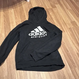 Svart Adidas hoodie med stor logga - Svart hoodie från Adidas med stor vit logga och texten 'ATHLETICS' på bröstet. Tröjan har huva och en klassisk känguruficka framtill. Perfekt för en sportig och avslappnad stil.