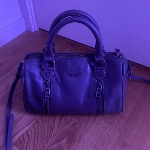 Svart Zadig & Voltaire handväska ”Sunny”  - (Pris går att diskuteras) Cool grå ”sunny” handväska från Zadig & Voltaire i mjukt skinn. Väskan har två rejäla handtag, avtagbar axelrem och snygga dragkedjedetaljer framtill. Medium storlek för allt du behöver ha med dig. Inte mini eller liten. 