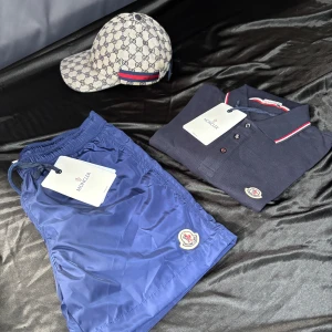 Blå Moncler badshorts med snörning - Snygga marinblå badshorts från Moncler med klassisk logga på benet. Shortsen har elastisk midja med snörning och praktisk bakficka med dragkedja. Tillverkade i lätt syntetmaterial som torkar snabbt – perfekta för stranden eller poolen. Shortsen har även NFC scan.