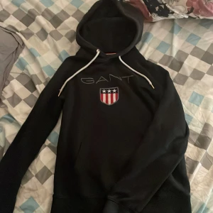 Svart hoodie från GANT med logga - Svart hoodie från GANT med vit snörning och stor broderad logga på bröstet. Klassisk huva och lång ärm, perfekt för en chill och sportig look. Tillverkad i mjuk bomullsmix som känns skön mot huden.