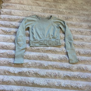 Ljusgrå croppad tröja från SOC - Snygg ljusgrå/beige croppad tröja från SOC med långa ärmar och diskret logga längs midjan. Tröjan är ribbad och har en tight passform, perfekt för träning eller chill. Mjukt och stretchigt material som sitter skönt på kroppen.