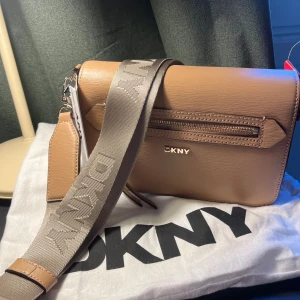 Beige axelväska från DKNY - Säljer min HELT nya väska jag fått i present som jag vet inte kommer gå till användning, då jag har en massa andra väskor. Ordinariepris 2899 kronor. Snygg beige axelväska från DKNY med bred, justerbar rem med logga. Väskan har ett stilrent, rektangulärt format, dragkedjeficka framtill och detaljer i guld. Tillverkad i skinnliknande material och perfekt för att bära det viktigaste. OBS jag kan tänka mig sänka priset vid snabb affär 