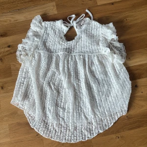 Vit spetsblus med volang från Nelly - vit blus från Nelly med volangärmar och spetsdetaljer. Blusen har knytning i nacken, luftig passform.