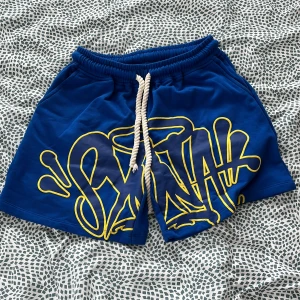 Blåa shorts från Syna world - Snygga blå shorts från Syna world med stor gul graffiti-print framtill. De har elastisk midja med vit snörning och är tillverkade i mjuk bomull. Perfekta för chill dagar eller när du vill sticka ut lite extra. Alla accessoarer finns, Påse, nyckel ring 