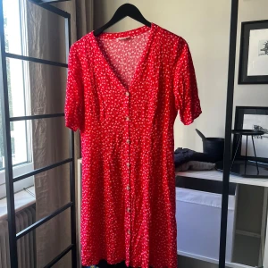 Röd blommig kortklänning från Monki - Superfin röd kortklänning från Monki med vita små blommor, v-ringning och knappar hela vägen framtill. Klänningen har korta ärmar och en lätt, flowy passform som känns både söt och chill. Perfekt för varma dagar när du vill sticka ut lite extra.
