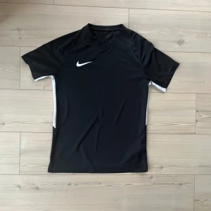 Svart Nike Dri-FIT fotbollströja - Svart fotbollströja från Nike med vit swoosh-logga på bröstet och vita detaljer längs sidorna. Tillverkad i lätt och ventilerande Dri-FIT-material som håller dig torr under matchen. Klassisk passform med korta ärmar och rund hals.