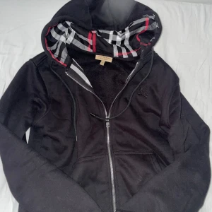 Svart Burberry hoodie med dragkedja - Svart hoodie från Burberry med klassiskt rutigt foder i huvan och broderad logga på bröstet. Jackan har dragkedja framtill, snörning i huvan och kängurufickor. Perfekt för dig som vill ha en stilren och ikonisk streetlook.