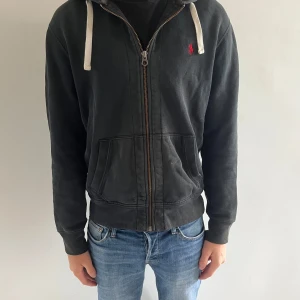 Svart hoodie från Ralph Lauren - Svart hoodie från Ralph Lauren med dragkedja, huva med vita snören och klassisk röd logga på bröstet. Tröjan har känguruficka framtill och ribbade muddar vid ärmslut och nederkant. Perfekt för en chill och avslappnad stil.